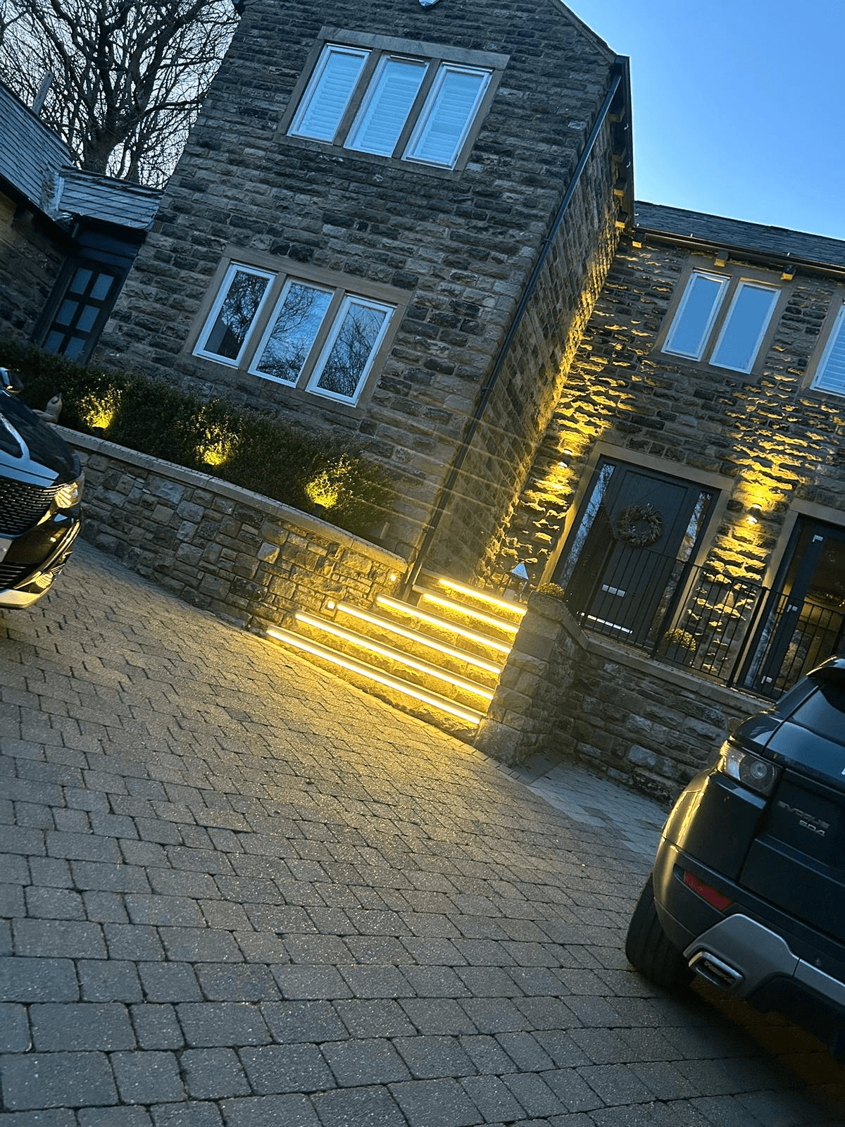 Stone house exterior with warm uplighting on steps and driveway area at dusk